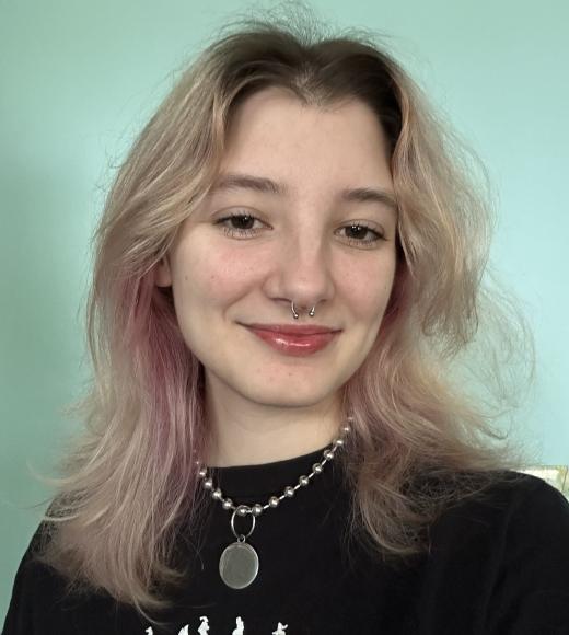 Smiling person with wavy blonde hair and a septum piercing, against a light teal background.