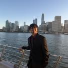 Young man in dark suit leaning on boat railing, city skyline and water at sunset.